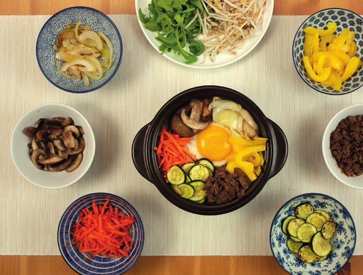 Angerichtetes Bibimbap im Tontopf mit Spiegelei und Gemüse, ringsum Schälchen, Rucola und Sprossen