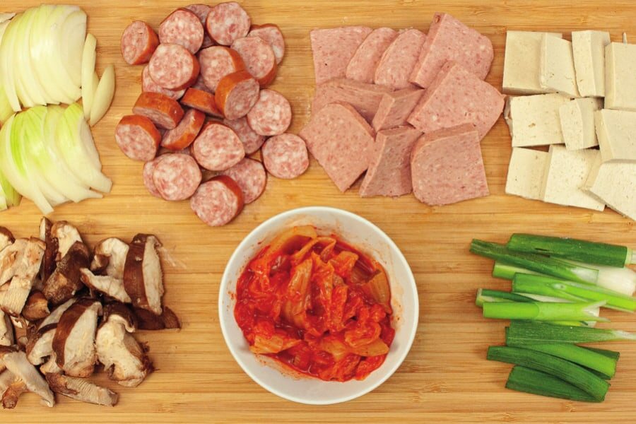 Geschnittene Zwiebel, Würstchen, Frühstücksfleisch, Tofu, Shiitake, Frühlingszwiebeln und eine Schale Kimchi auf einem Brett