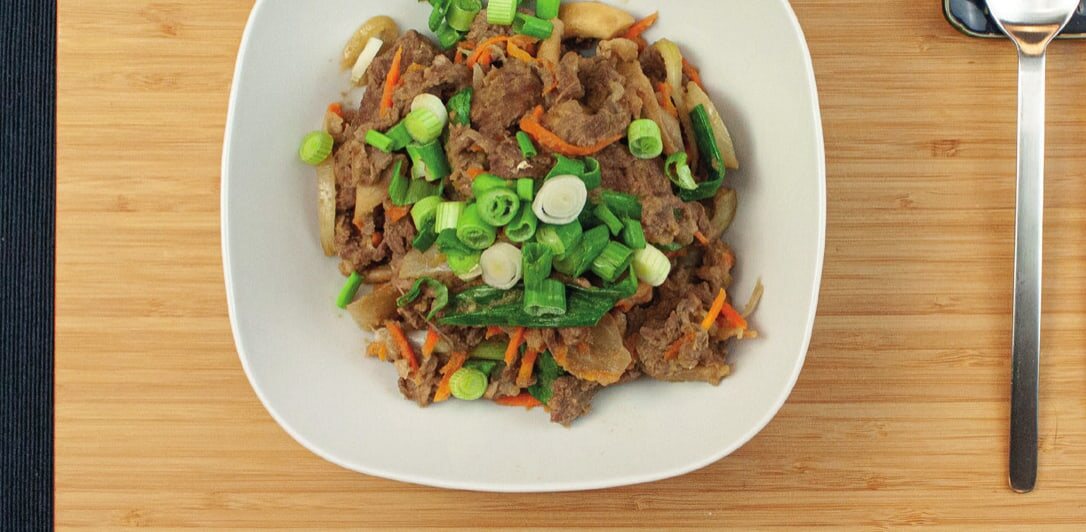 Bulgogi mit Karottenstreifen und Frühlingszwiebelringen in einer weißen Schale, daneben Löffel und Stäbchen