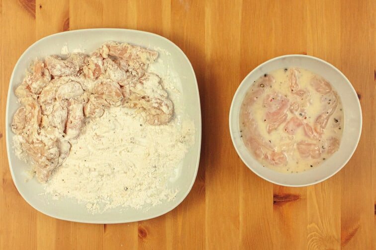 Panierte Hähnchenstücke auf einem Teller mit Mehlmischung, daneben die Schüssel mit Teig und Fleisch