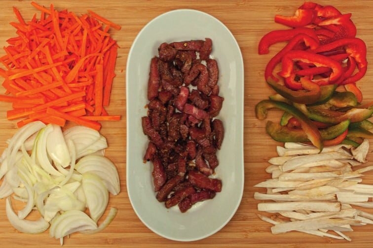 Karotten-, Zwiebel-, Paprika- und Pilzstreifen auf einem Brett, in der Mitte mariniertes Rindfleisch