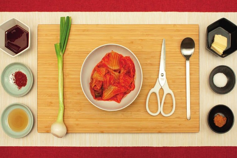 Kimchi in einer Schale, Frühlingszwiebel und Schere auf einem Brett, rundherum Schälchen mit Sojasoße, Chiliflocken, Sesamöl, Butter, Zucker und Doenjang