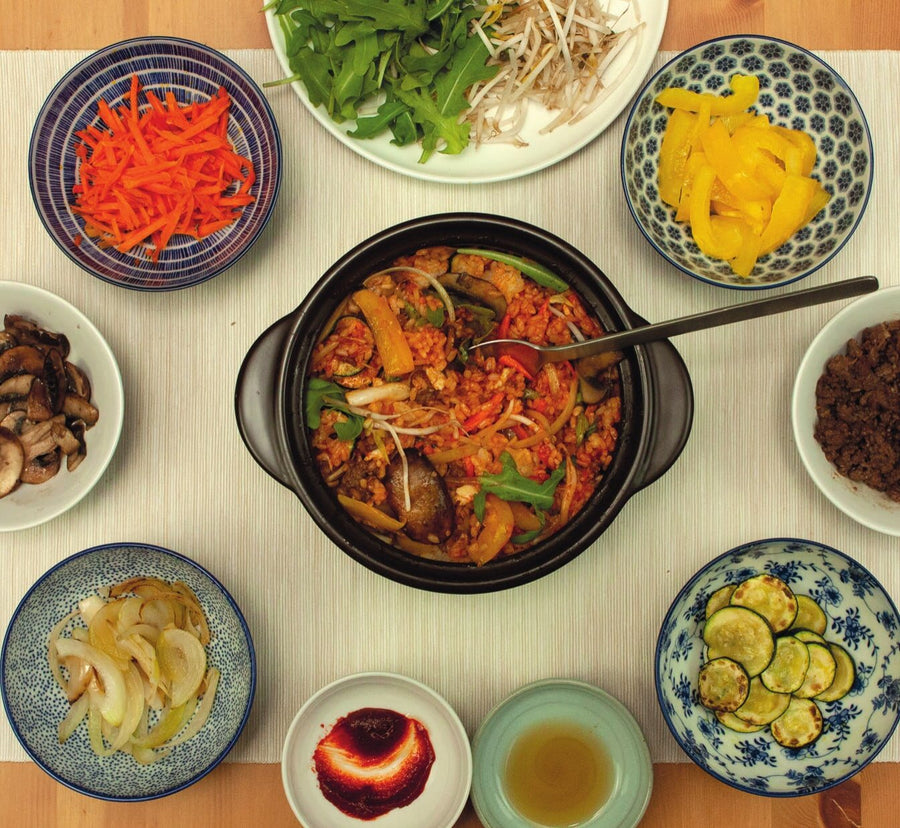Durchgemischtes Bibimbap im Tontopf mit Löffel, ringsum Schälchen mit Gemüse, Gochujang und Sesamöl