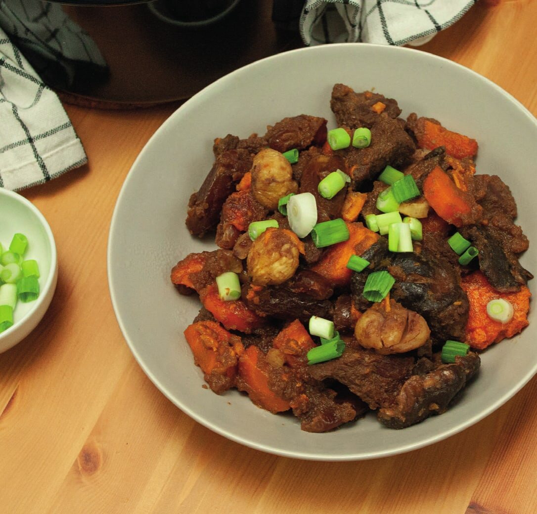 Galbi-Jjim auf einem Teller mit Karotte, Süßkartoffel, Maronen und Frühlingszwiebeln, dahinter Topf und eine Schale Reis