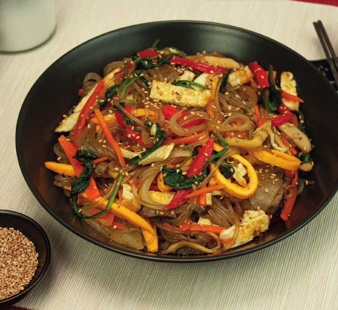 Japchae in einer dunklen Schale mit Paprika, Spinat, Eistreifen und Sesam