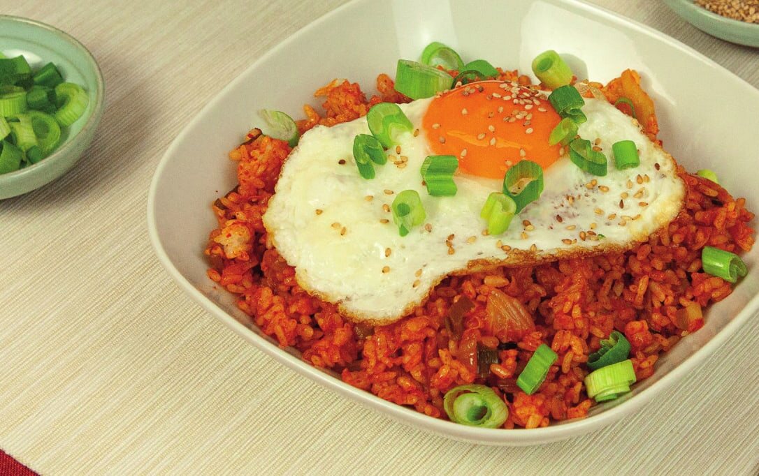 Gebratener Kimchi-Reis mit Spiegelei, Frühlingszwiebeln und Sesam in einer hellen Schale, daneben ein Löffel