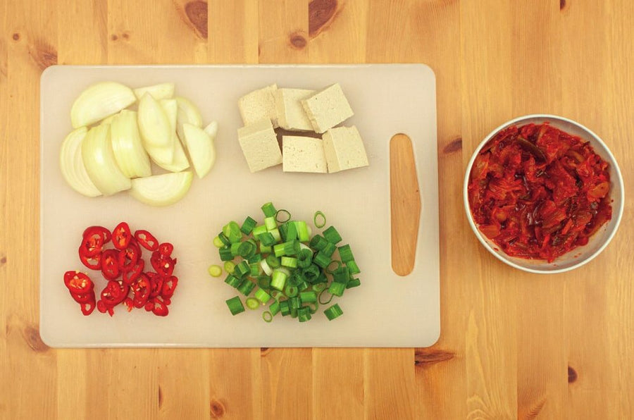 Geschnittene Zwiebel, Tofu, Chili und Frühlingszwiebel auf einem Brett, daneben eine Schale Kimchi